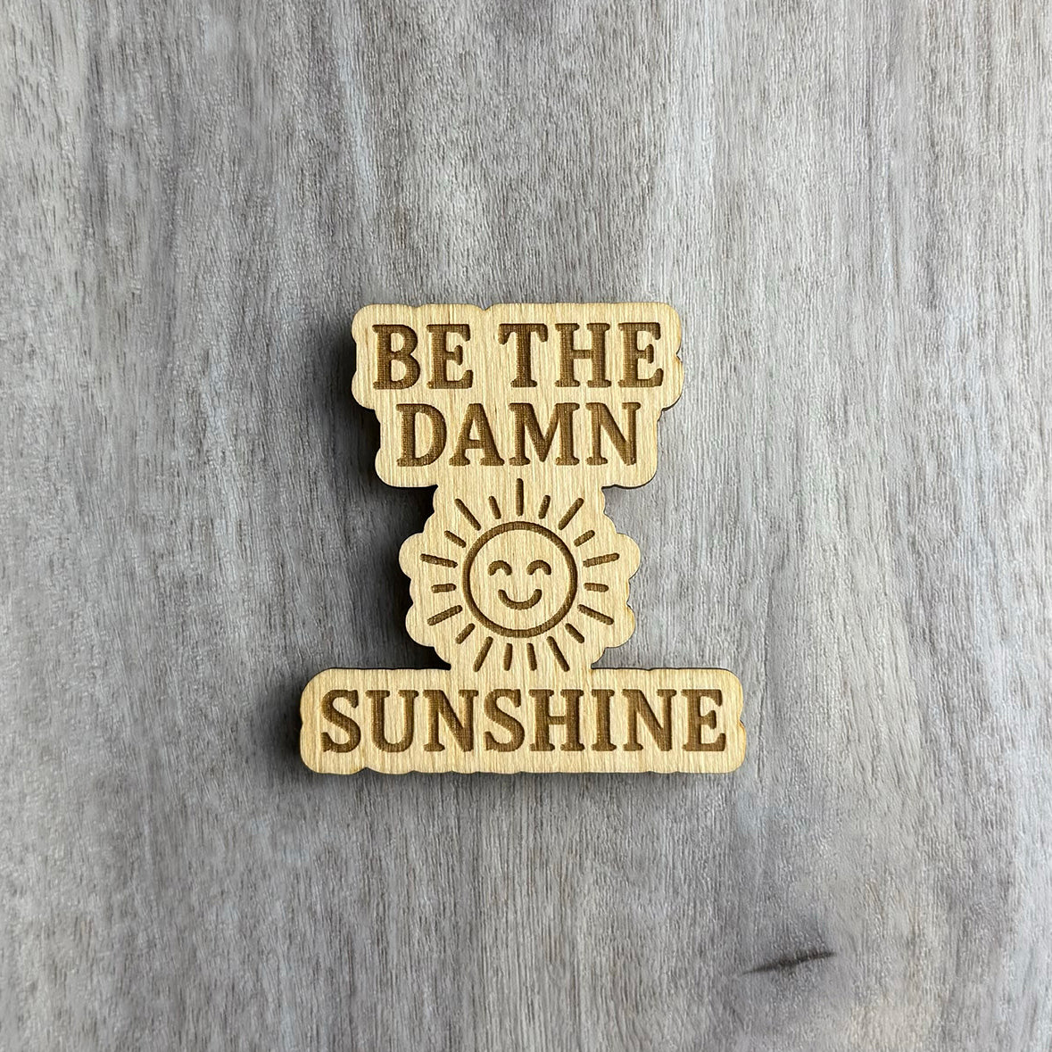 Laser-cut wooden magnet with the phrase Be the Damn Sunshine engraved in bold lettering, separated by a smiling sun illustration in the center, placed on a gray wood background.