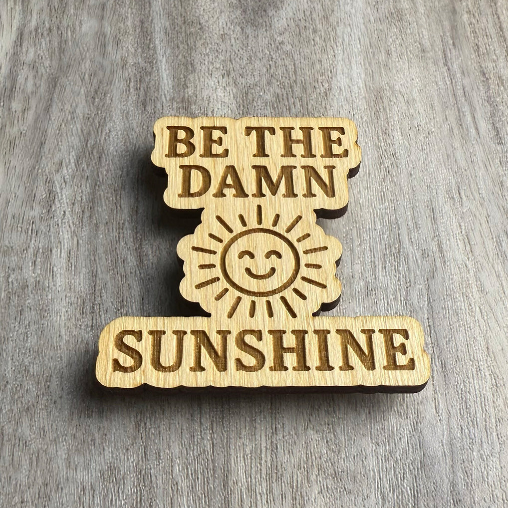 Angled view of a laser-cut wooden magnet featuring the engraved phrase Be the Damn Sunshine with a happy sun design in the middle, showing the depth of the cut and thickness of the wood, resting on a gray wood surface.