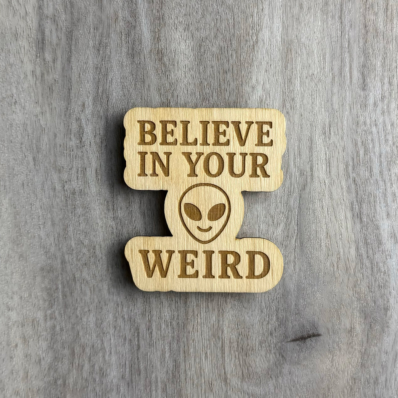 Laser-cut wooden magnet engraved with the phrase Believe in Your Weird, featuring a stylized alien face in the center between the text, displayed on a gray wood background.