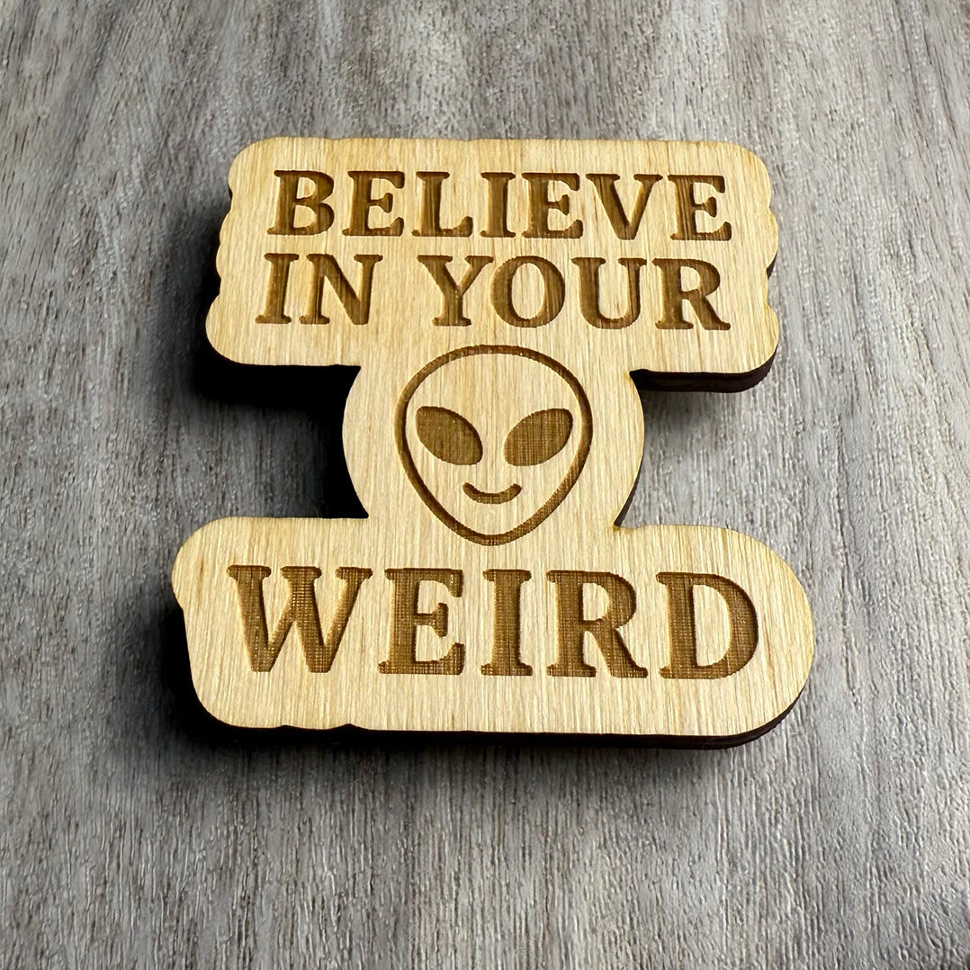 Angled view of a laser-cut wooden magnet engraved with the phrase Believe in Your Weird, featuring a bold alien face in the center, highlighting the depth of the engraving and the layered wood construction on a gray wood surface.