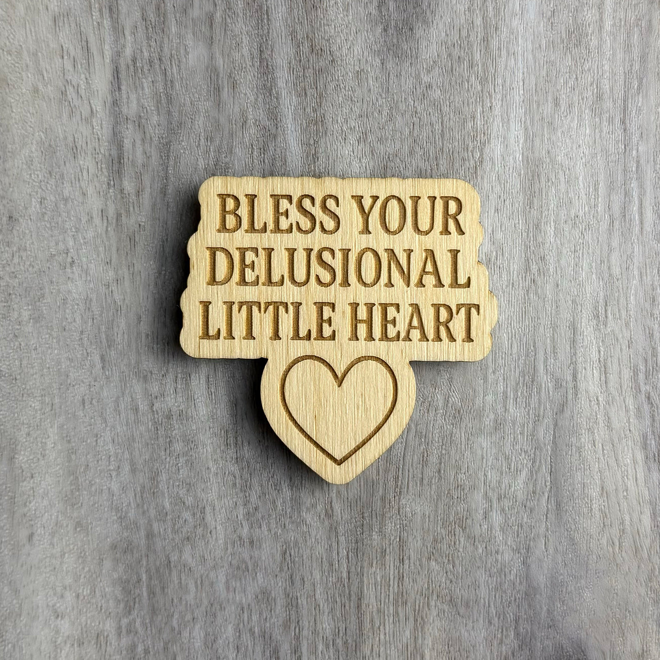 Laser-cut wooden magnet with the phrase Bless Your Delusional Little Heart engraved in bold text above a simple outlined heart shape, set against a gray wood background.