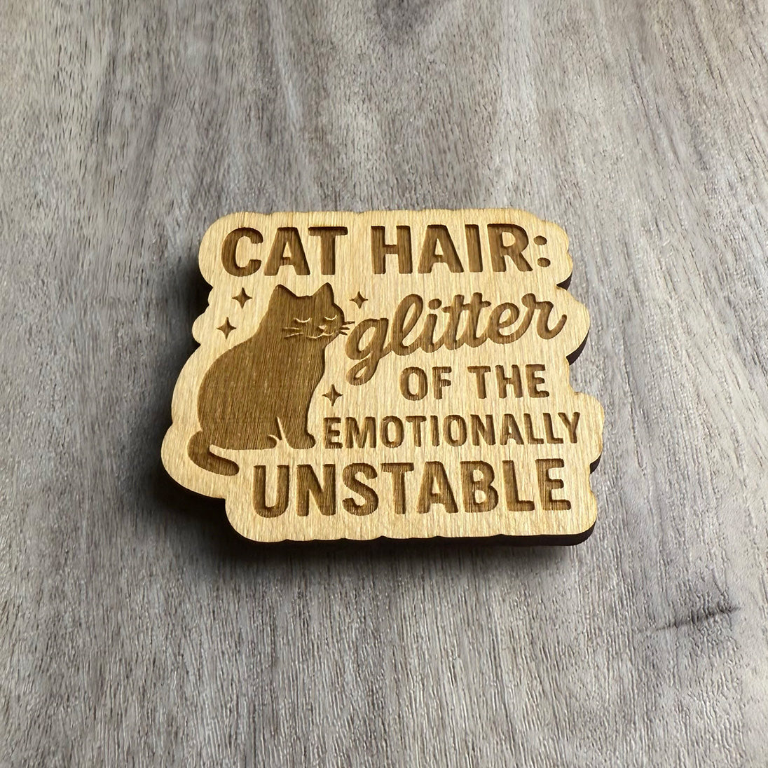 Angled view of a laser-cut wooden magnet with the engraved phrase Cat Hair: Glitter of the Emotionally Unstable and a cat with sparkles, showing the engraved depth and layered wood on a gray surface.