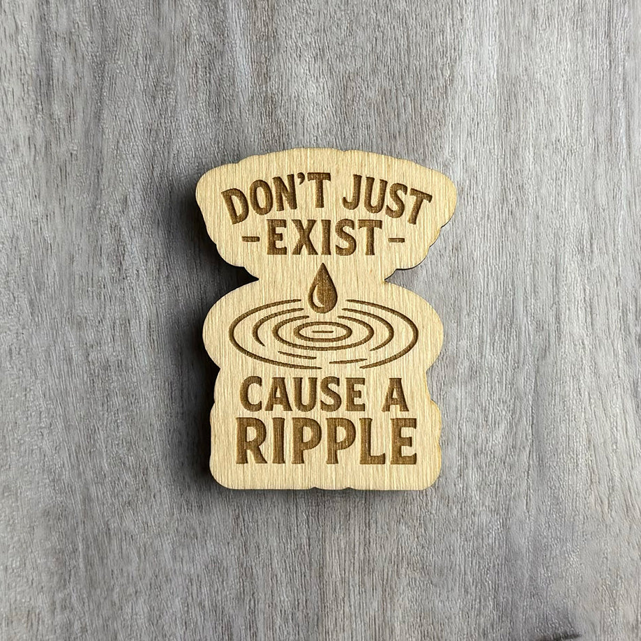 Laser-cut wooden magnet engraved with the phrase Don’t Just Exist — Cause a Ripple, featuring a water droplet above concentric ripple rings in the center, set on a gray wood background.