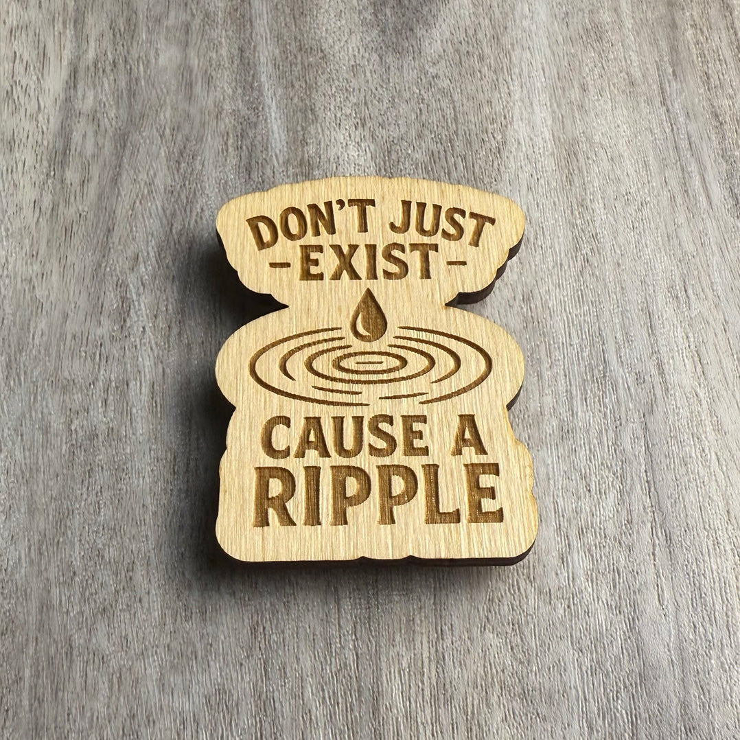 Angled view of a laser-cut wooden magnet displaying the message Don’t Just Exist — Cause a Ripple, with a droplet and ripple illustration in the center, showing engraving depth and material thickness on a gray surface.