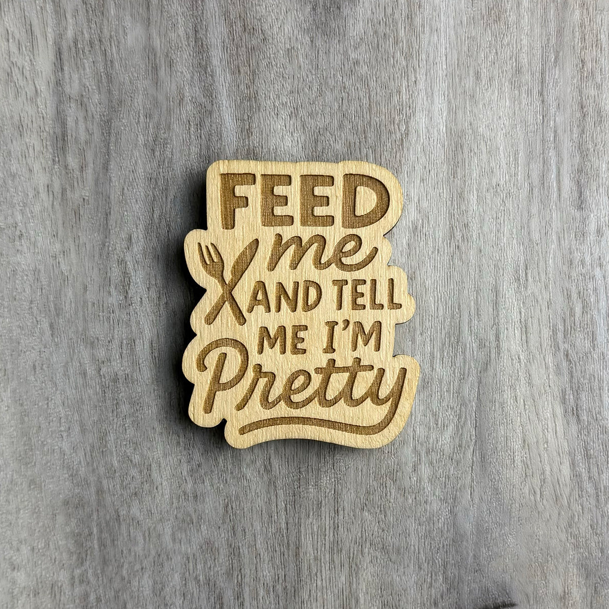 Laser-cut wooden magnet engraved with the phrase Feed Me and Tell Me I’m Pretty, featuring a fork and knife crossed in the center, styled with mixed fonts and placed on a gray wood background.