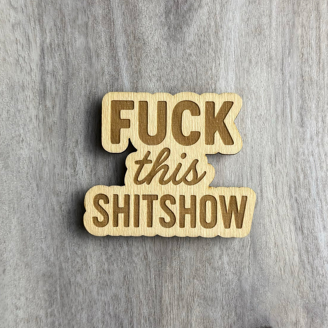 Laser-cut wooden magnet engraved with the phrase Fuck This Shitshow in a mix of bold and script fonts, placed on a gray wood background.