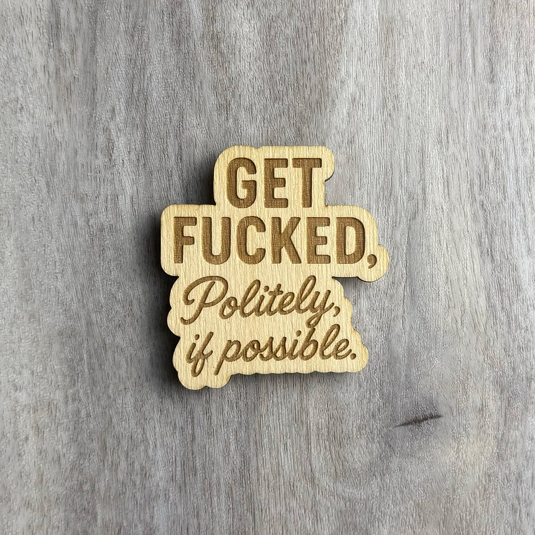 Laser-cut wooden magnet engraved with the phrase Get Fucked, Politely, if possible, using a combination of bold block lettering and cursive script, displayed on a gray wood background.