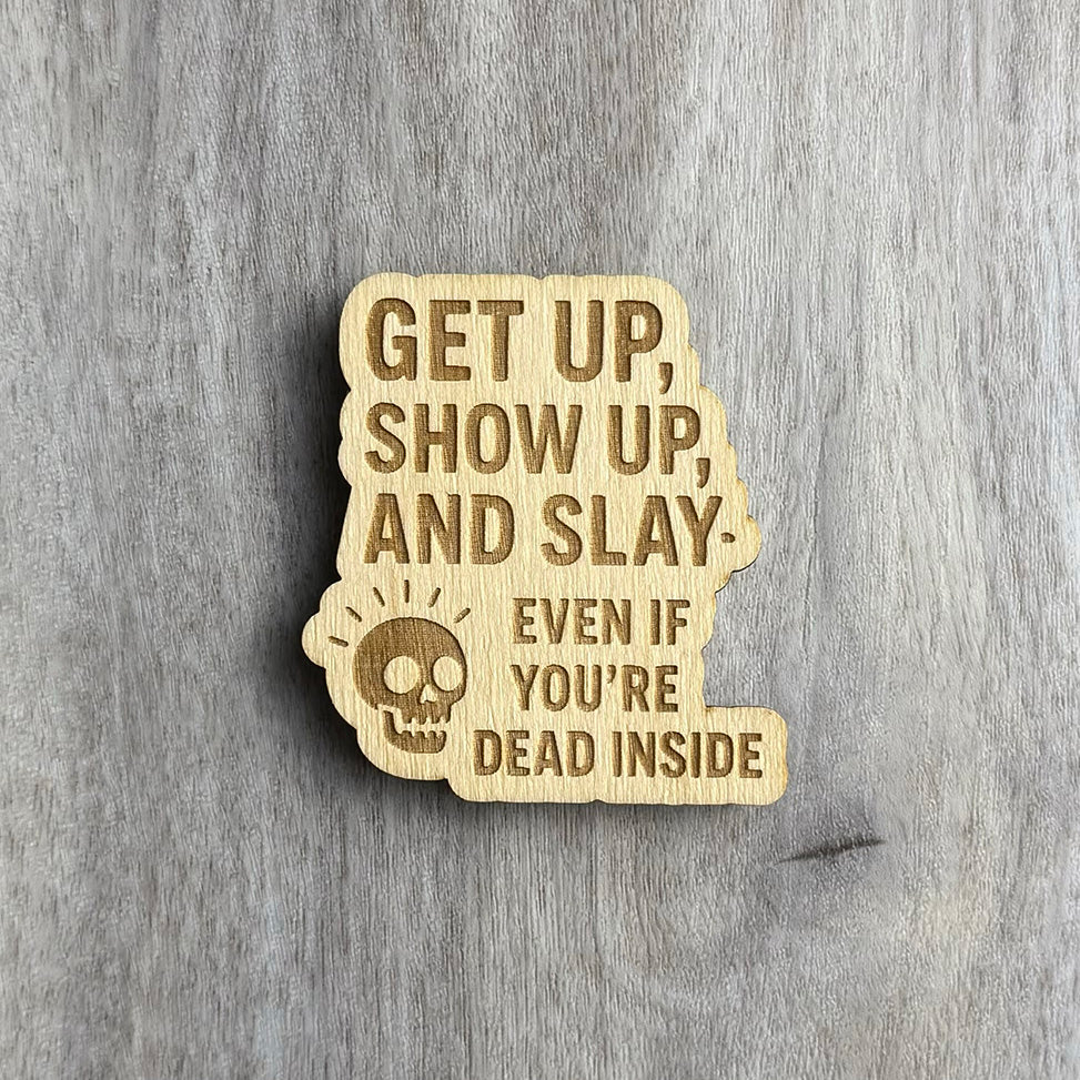 Laser-cut wooden magnet engraved with the phrase Get Up, Show Up, and Slay — Even If You're Dead Inside, featuring a small skull illustration with radiating lines, displayed on a gray wood background.