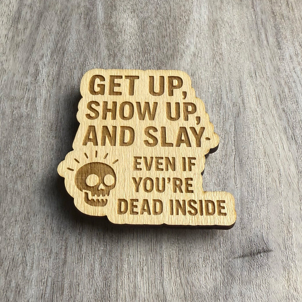 Angled view of a laser-cut wooden magnet with the phrase Get Up, Show Up, and Slay — Even If You're Dead Inside and a skull icon, highlighting the engraving depth and thickness of the wooden piece on a gray surface.