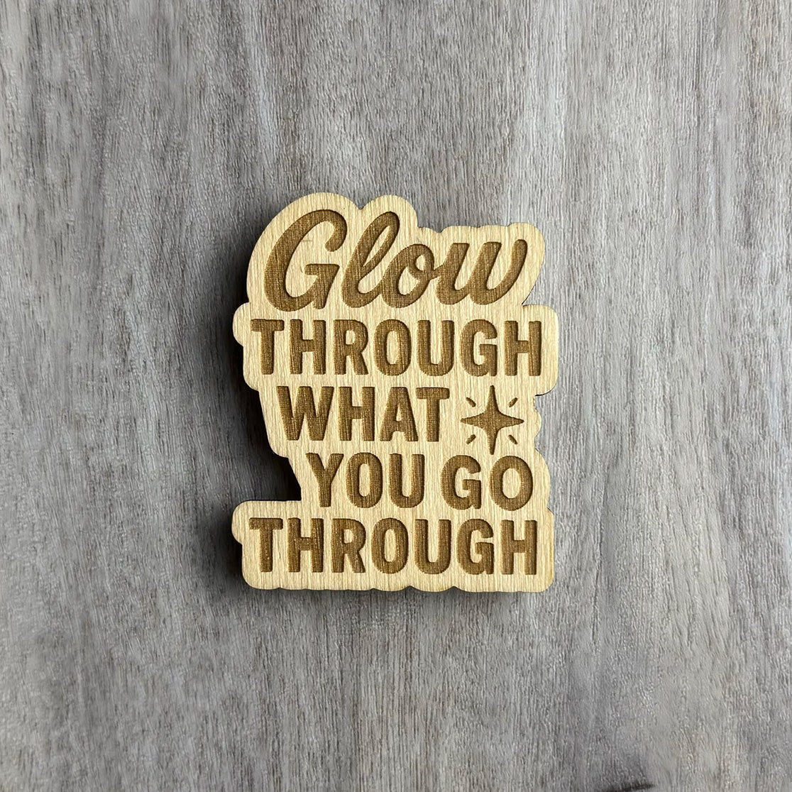 Laser-cut wooden magnet engraved with the motivational phrase Glow Through What You Go Through, featuring a small starburst accent, displayed on a gray wood background.