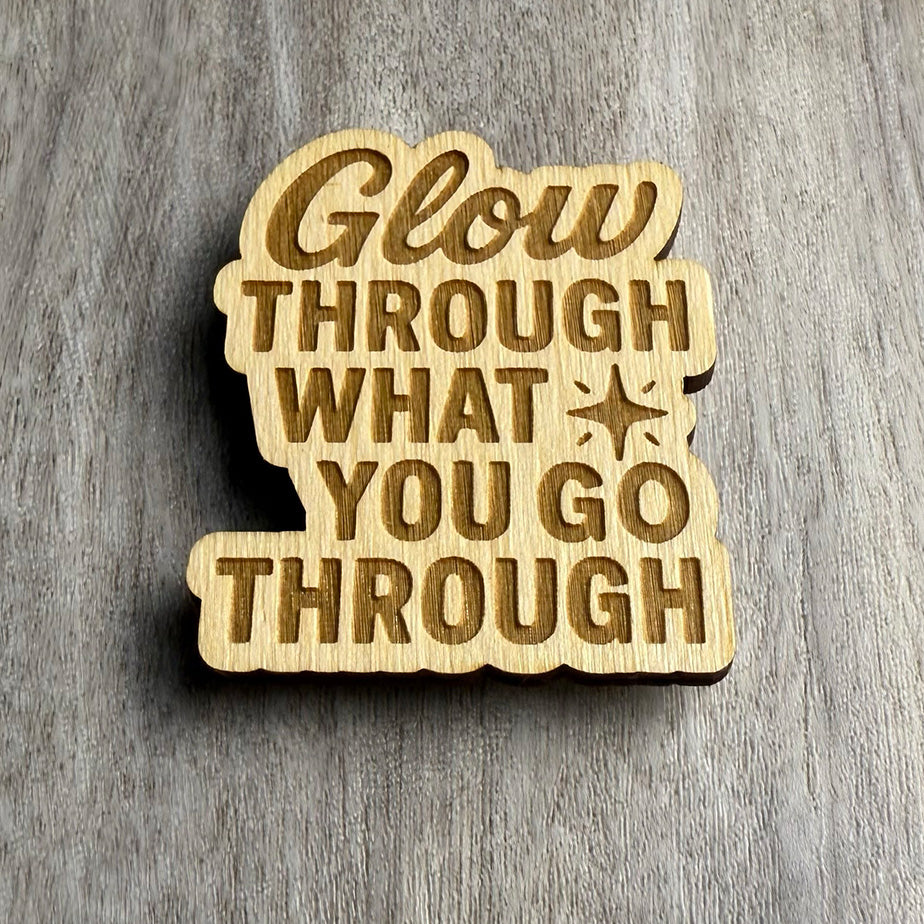 Angled view of a laser-cut wooden magnet with the phrase Glow Through What You Go Through and a starburst symbol, showcasing the engraved detail and thickness of the wooden piece on a gray surface.