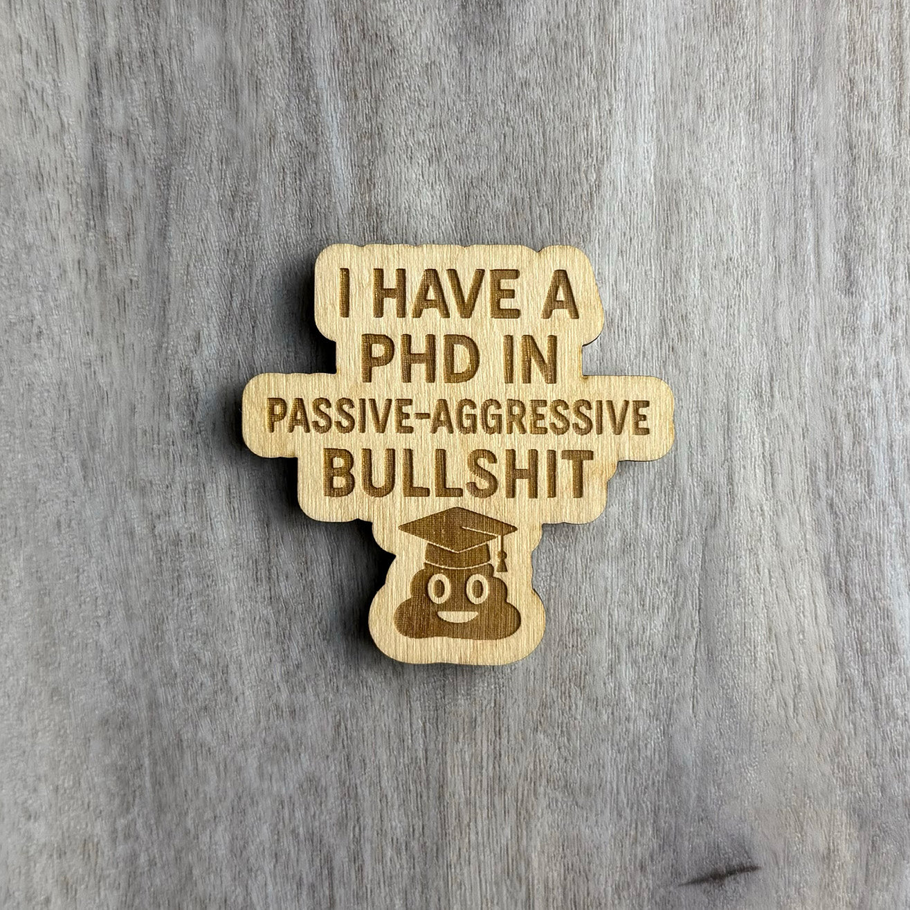 Laser-cut wooden magnet engraved with the phrase I Have a PhD in Passive-Aggressive Bullshit, featuring a smiling poop emoji wearing a graduation cap at the bottom, displayed on a gray wood background.