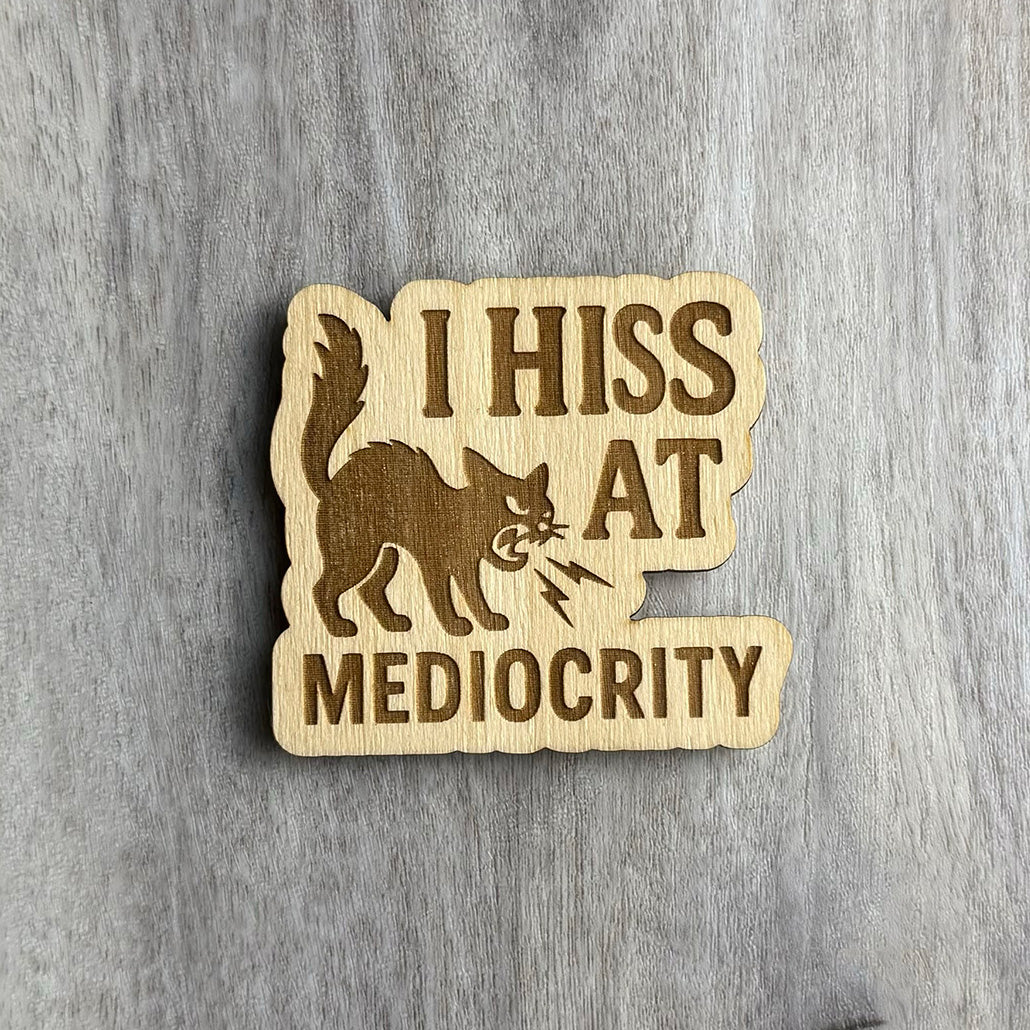 Laser-cut wooden magnet engraved with the phrase I Hiss at Mediocrity, featuring an angry, arched-back cat hissing with lightning bolts coming from its mouth, set against a gray wood background.