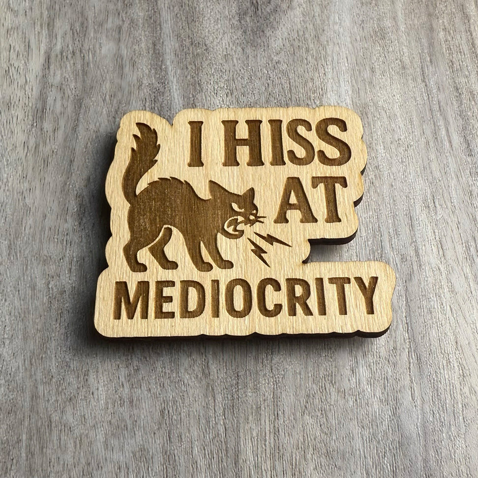 Angled view of a laser-cut wooden magnet showing the phrase I Hiss at Mediocrity and a fierce hissing cat illustration, highlighting engraving depth and wood thickness on a gray surface.