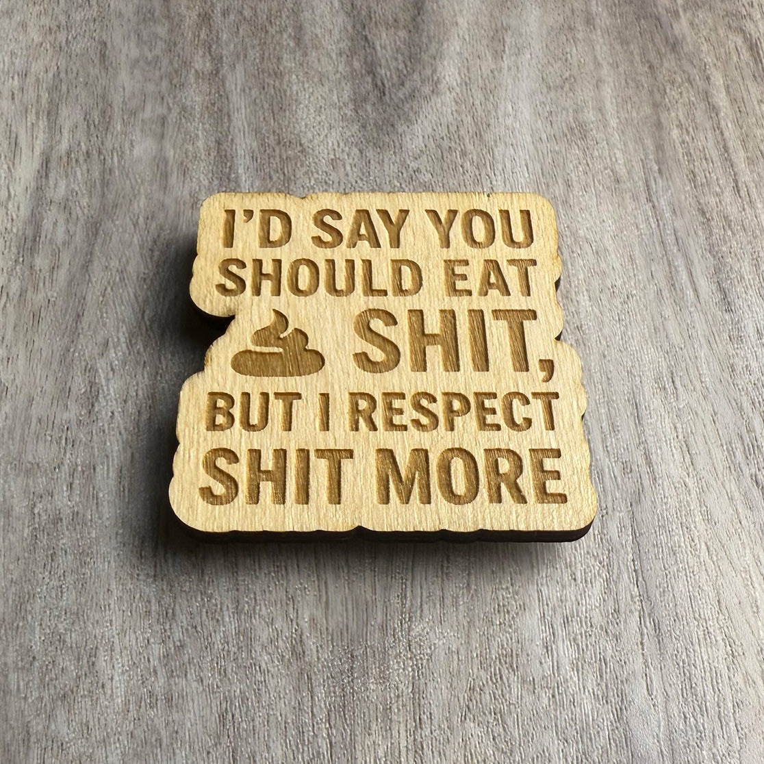Angled view of a wooden magnet engraved with the phrase “I’D SAY YOU SHOULD EAT SHIT, BUT I RESPECT SHIT MORE.” The lighting highlights the engraved depth and natural texture of the wood.