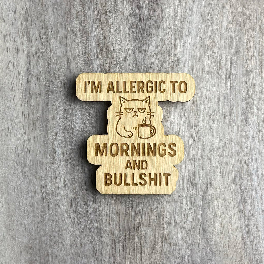 Laser-engraved wooden magnet featuring the phrase “I’M ALLERGIC TO MORNINGS AND BULLSHIT” in bold lettering. At the center is a grumpy-looking cat holding a steaming coffee mug, engraved in line art. The magnet has a custom cutout shape following the design elements.