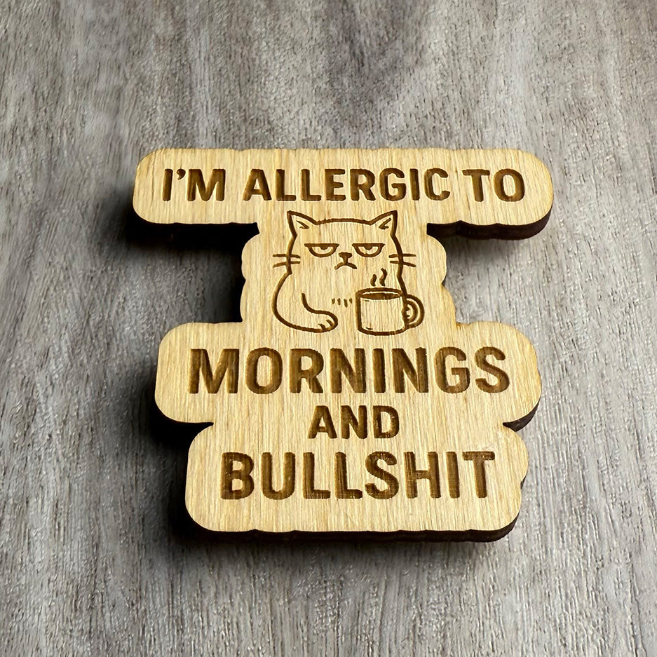 Angled view of the same engraved wooden magnet with the phrase “I’M ALLERGIC TO MORNINGS AND BULLSHIT” and a sleepy, annoyed cat holding a coffee mug. The engraved lines and wood grain are emphasized by side lighting.