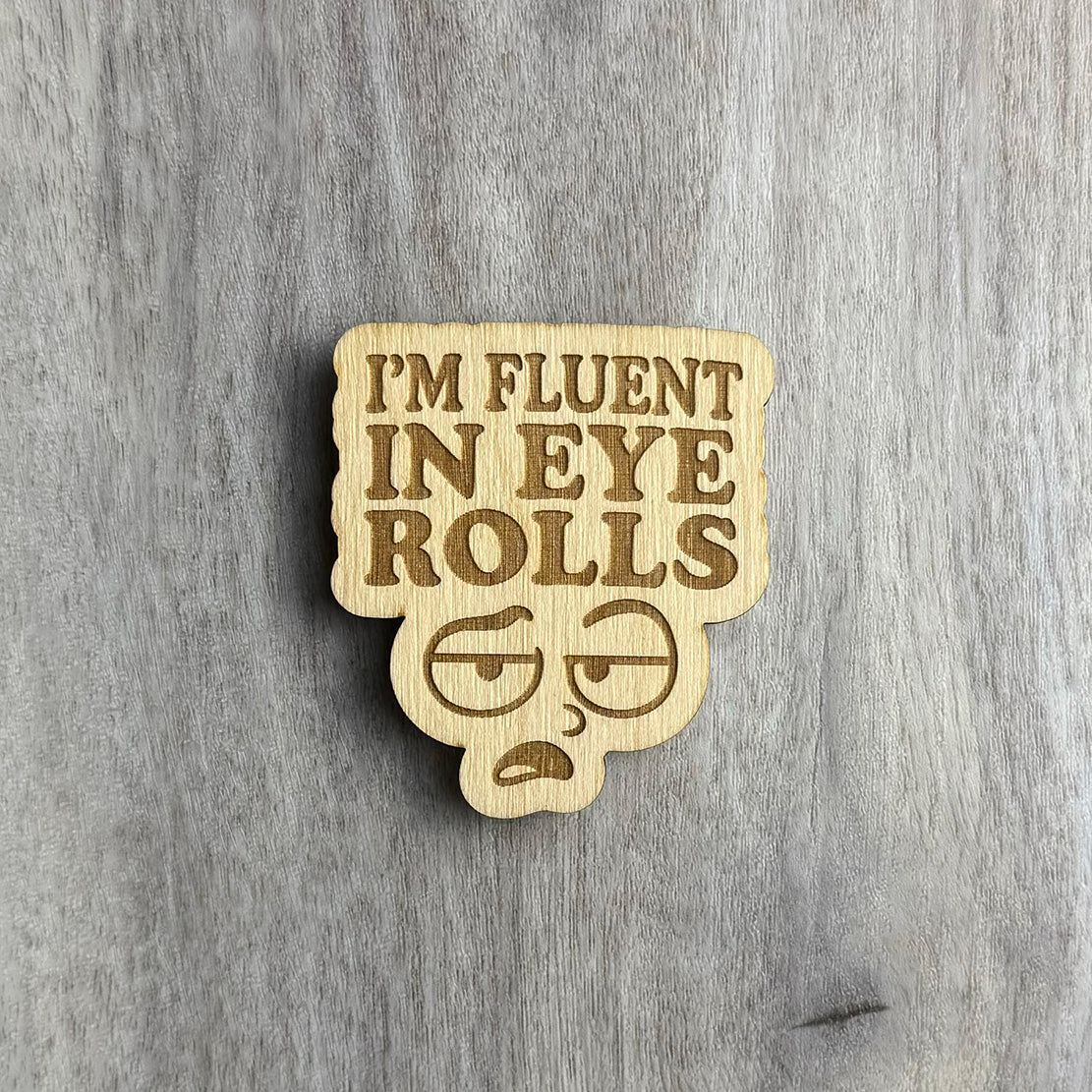 Laser-engraved wooden magnet with the phrase “I’M FLUENT IN EYE ROLLS” above a cartoon-style face featuring exaggerated eyes mid-roll, a furrowed brow, and a sarcastic expression. The magnet has a custom cutout shape that follows the contours of the text and face.