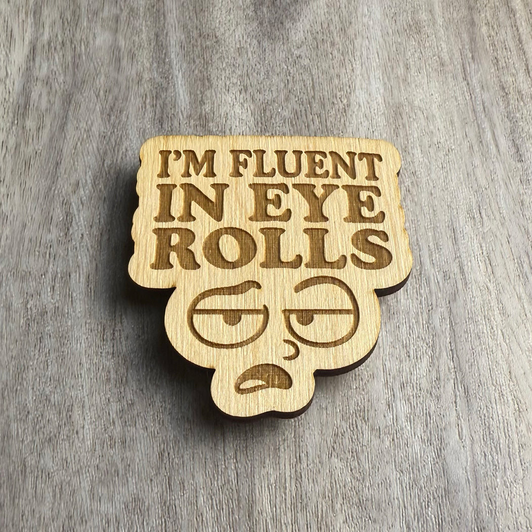 Angled view of the same engraved wooden magnet reading “I’M FLUENT IN EYE ROLLS,” with a deadpan cartoon face below the text. The lighting accentuates the engraved details and the layered wood texture.