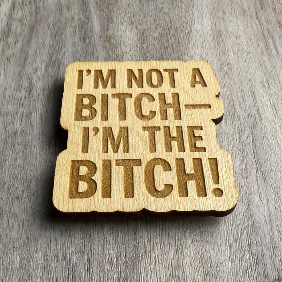 Angled view of a wooden magnet engraved with the phrase “I’M NOT A BITCH— I’M THE BITCH!” in bold uppercase letters. The lighting adds dimension to the engraved text and highlights the natural wood texture.