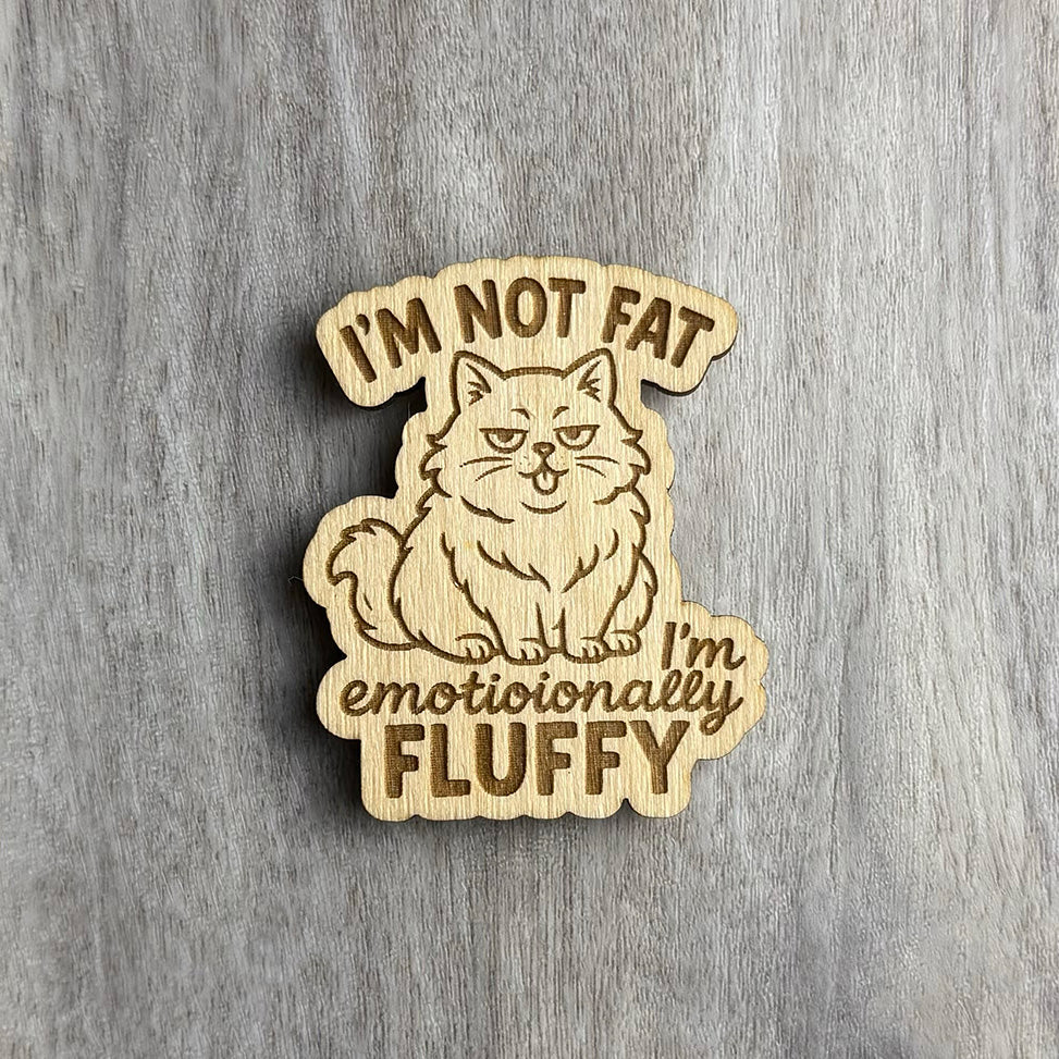 Laser-engraved wooden magnet with the phrase “I’M NOT FAT, I’m emotionally FLUFFY.” The design features a smug, fluffy cat sticking out its tongue, with a mix of script and bold fonts. The magnet shape follows the outline of the cat and text.