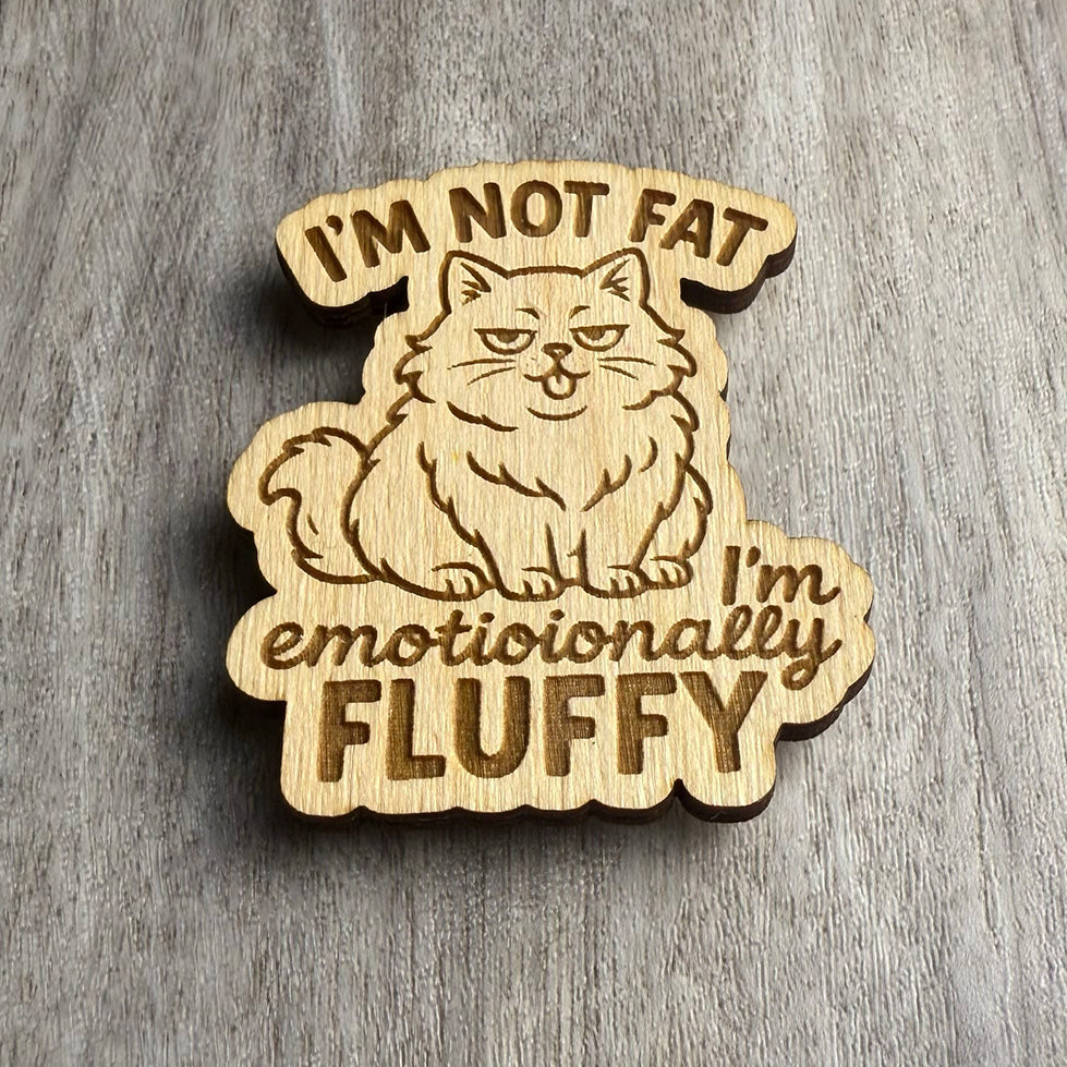 Angled view of the same engraved wooden magnet showing the phrase “I’M NOT FAT, I’m emotionally FLUFFY” alongside a plump, self-satisfied cat illustration. The engraving depth and natural wood grain are highlighted by directional lighting.