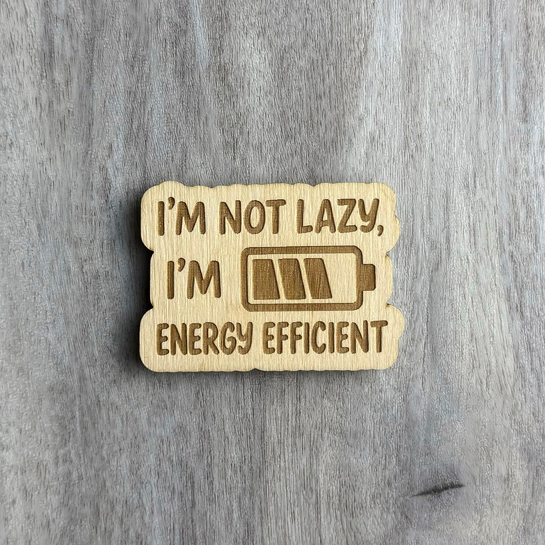 Laser-engraved wooden magnet featuring the phrase “I’M NOT LAZY, I’M ENERGY EFFICIENT” in a mix of bold and rounded font. A partially filled battery icon is engraved between the lines. The shape of the magnet follows a soft, bubbly border around the design.