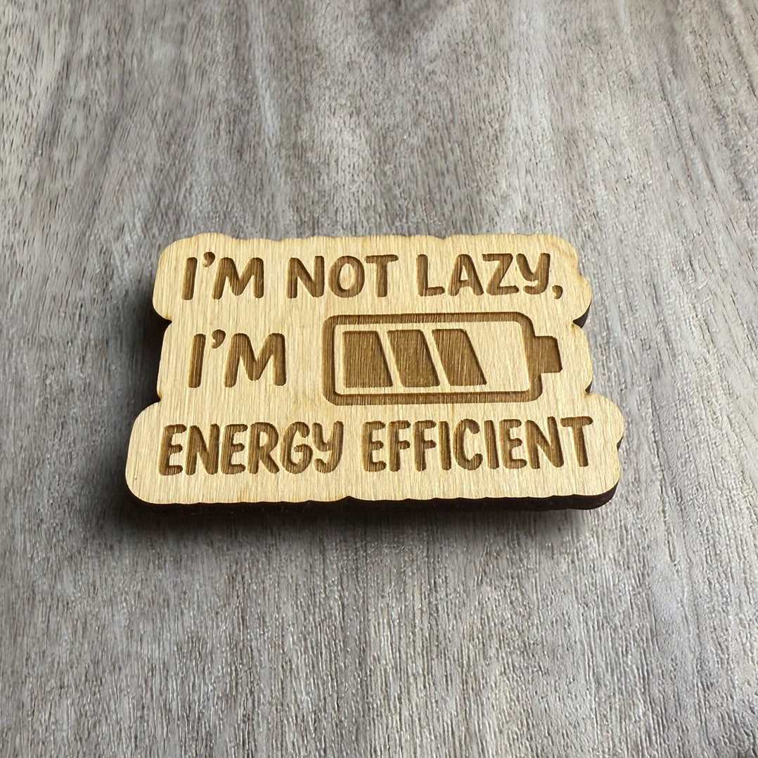 Angled view of the same engraved wooden magnet with the phrase “I’M NOT LAZY, I’M ENERGY EFFICIENT” and a stylized battery icon. Lighting highlights the engraved text and icon, emphasizing the natural wood texture and thickness.