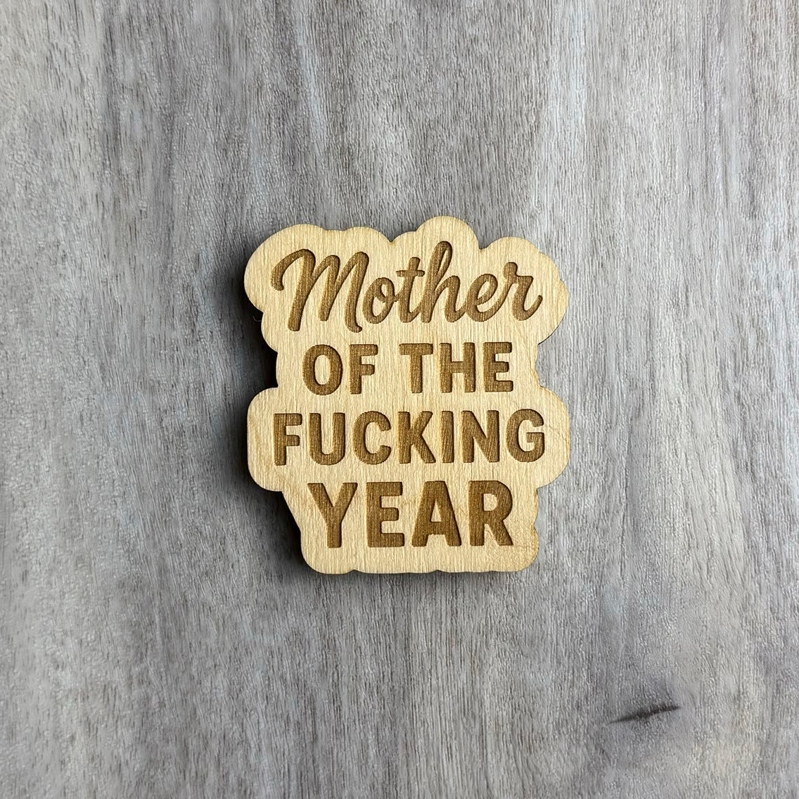 Laser-engraved wooden magnet with the phrase “Mother OF THE FUCKING YEAR.” The word “Mother” is in a cursive script, while the rest is in bold block letters. The magnet has a cloud-like, organic border shape that wraps around the stacked text.
