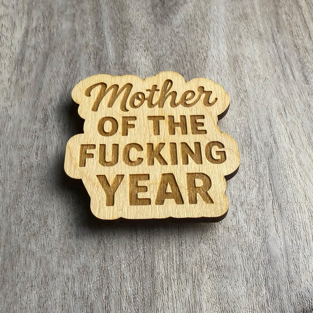 Angled view of the same engraved wooden magnet reading “Mother OF THE FUCKING YEAR,” combining elegant script and bold font styles. The lighting emphasizes the engraved depth and rounded edges of the custom cut shape.