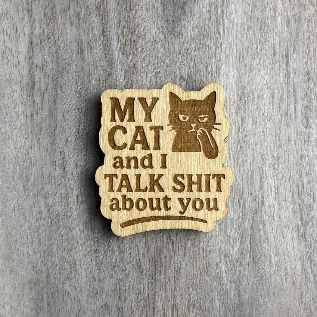 Laser-engraved wooden magnet with the phrase “MY CAT and I TALK SHIT about you.” The design includes a smug-looking cat with its paw to its mouth in a whispering pose. The magnet has a rounded rectangular shape with soft wave-like edges.