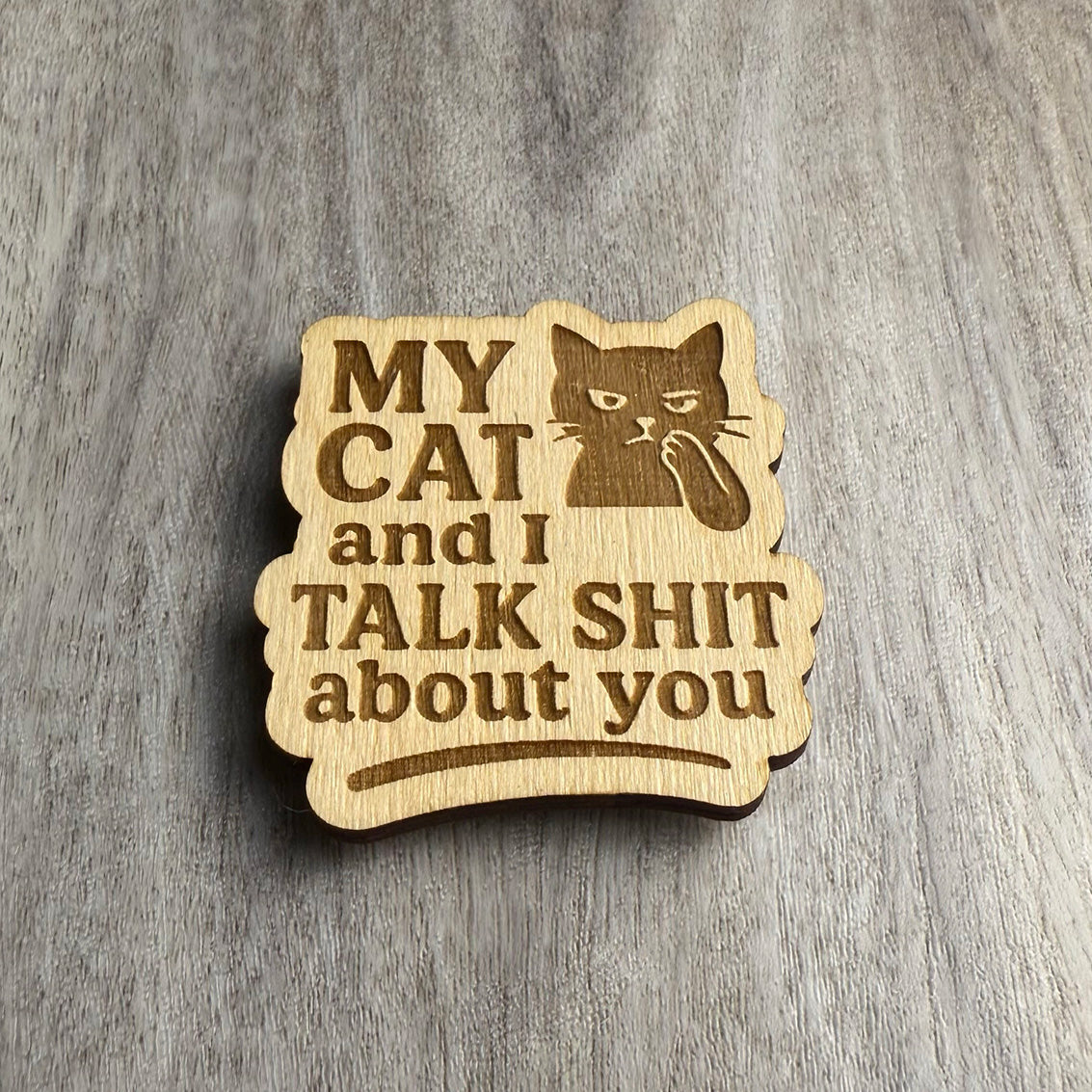 Angled view of the same engraved wooden magnet that says “MY CAT and I TALK SHIT about you,” featuring a gossipy cat illustration. Lighting enhances the engraved details and layered wood grain texture.