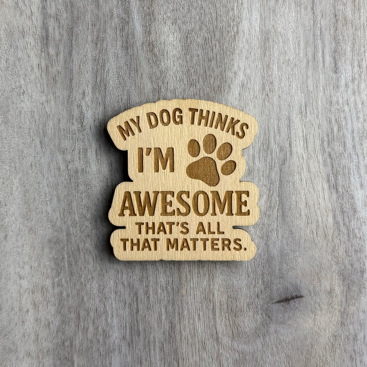 Laser-engraved wooden magnet with the phrase “MY DOG THINKS I’M AWESOME THAT’S ALL THAT MATTERS.” A paw print icon is engraved between the words “I’M” and “AWESOME.” The magnet has a stacked, curved shape with rounded, flowing edges.
