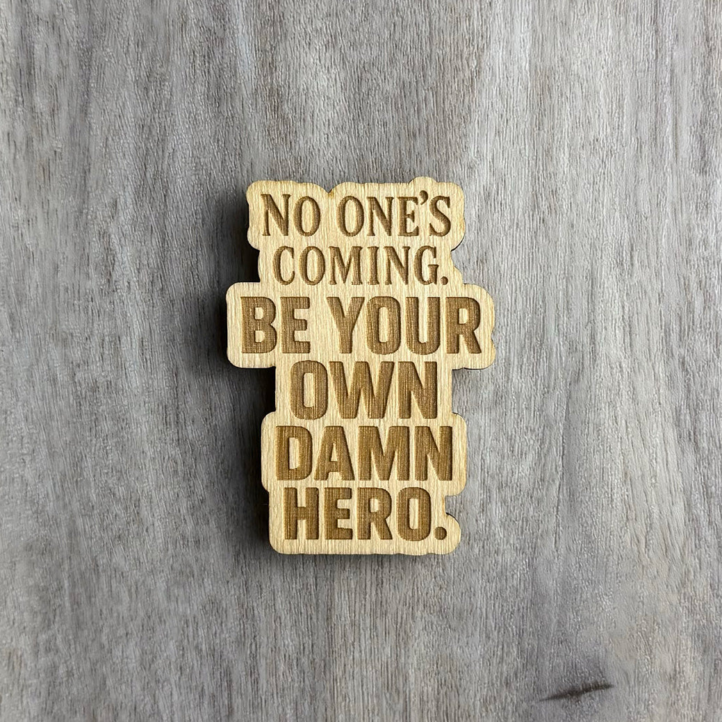 Laser-engraved wooden magnet with the empowering phrase “NO ONE’S COMING. BE YOUR OWN DAMN HERO.” The text is arranged in a tall stacked format using a mix of uppercase serif and bold block fonts. The shape of the magnet follows the rectangular structure of the layout.