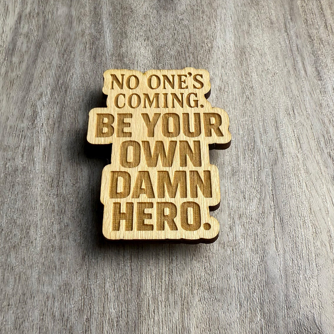 Angled view of the engraved wooden magnet that reads “NO ONE’S COMING. BE YOUR OWN DAMN HERO.” The lighting brings out the engraved depth and natural wood grain, adding emphasis to the bold typography.