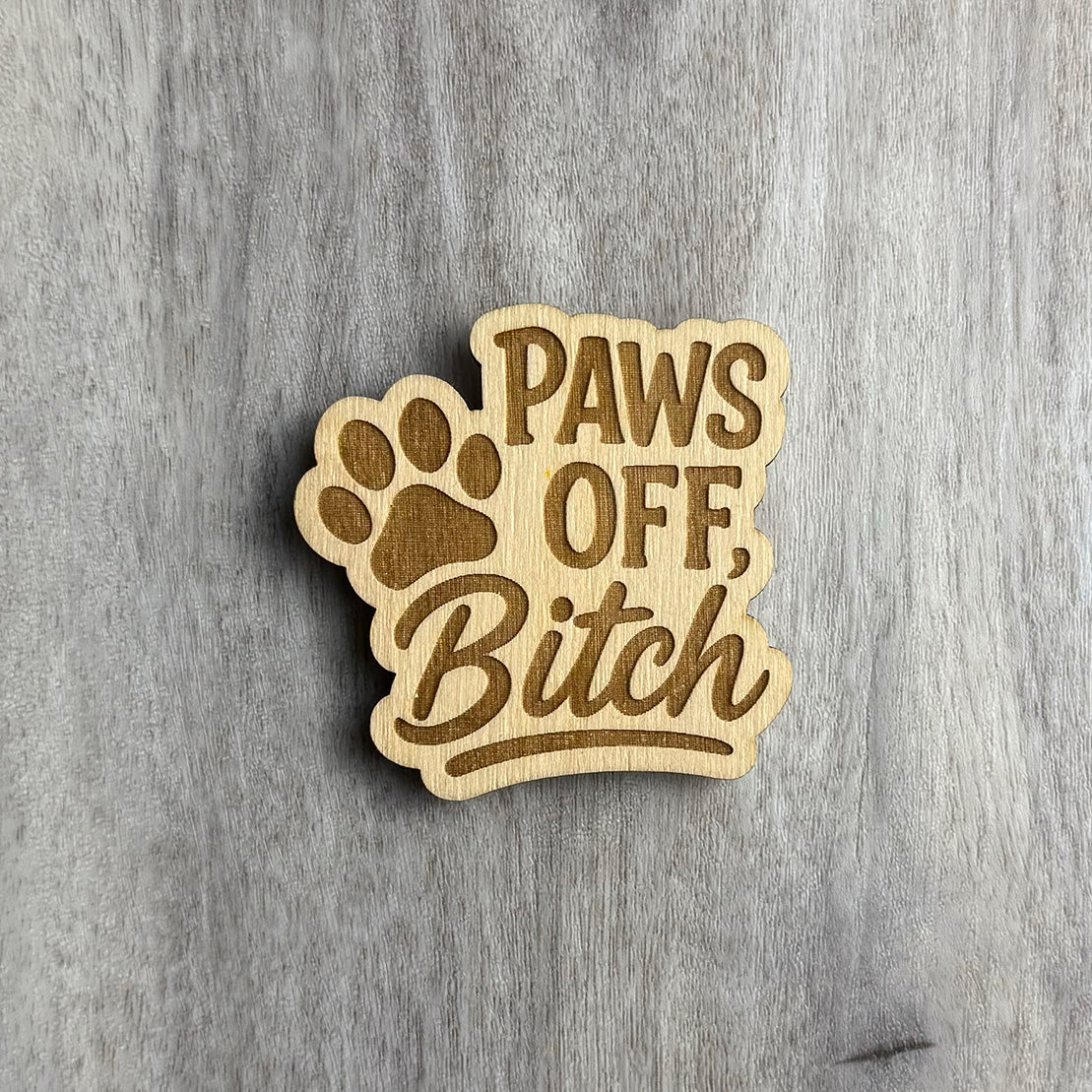 Laser-engraved wooden magnet with the phrase “PAWS OFF, Bitch” in a combination of bold uppercase and flowing script fonts. A large engraved paw print sits behind the word "PAWS," integrated into the design. The magnet has a playful cutout shape that follows the text and paw outline.