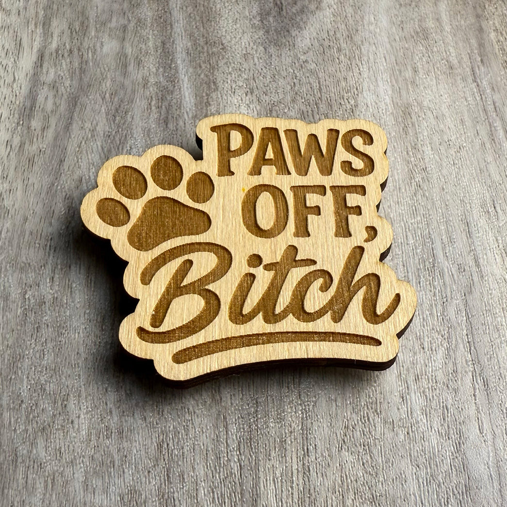 Angled view of the same engraved wooden magnet reading “PAWS OFF, Bitch,” with a bold paw print above the text. The lighting highlights the engraving depth and smooth grain of the wood, giving the design a strong, cheeky flair.