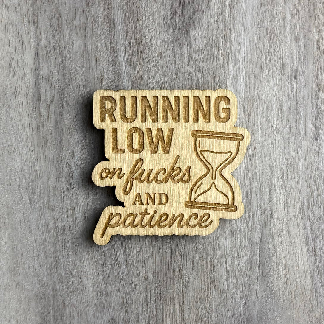 Wooden magnet engraved with the bold phrase "RUNNING LOW on fucks AND patience," featuring a mix of block and cursive fonts. An hourglass icon is engraved to the right, symbolizing time running out. The background is a light gray wood grain surface.