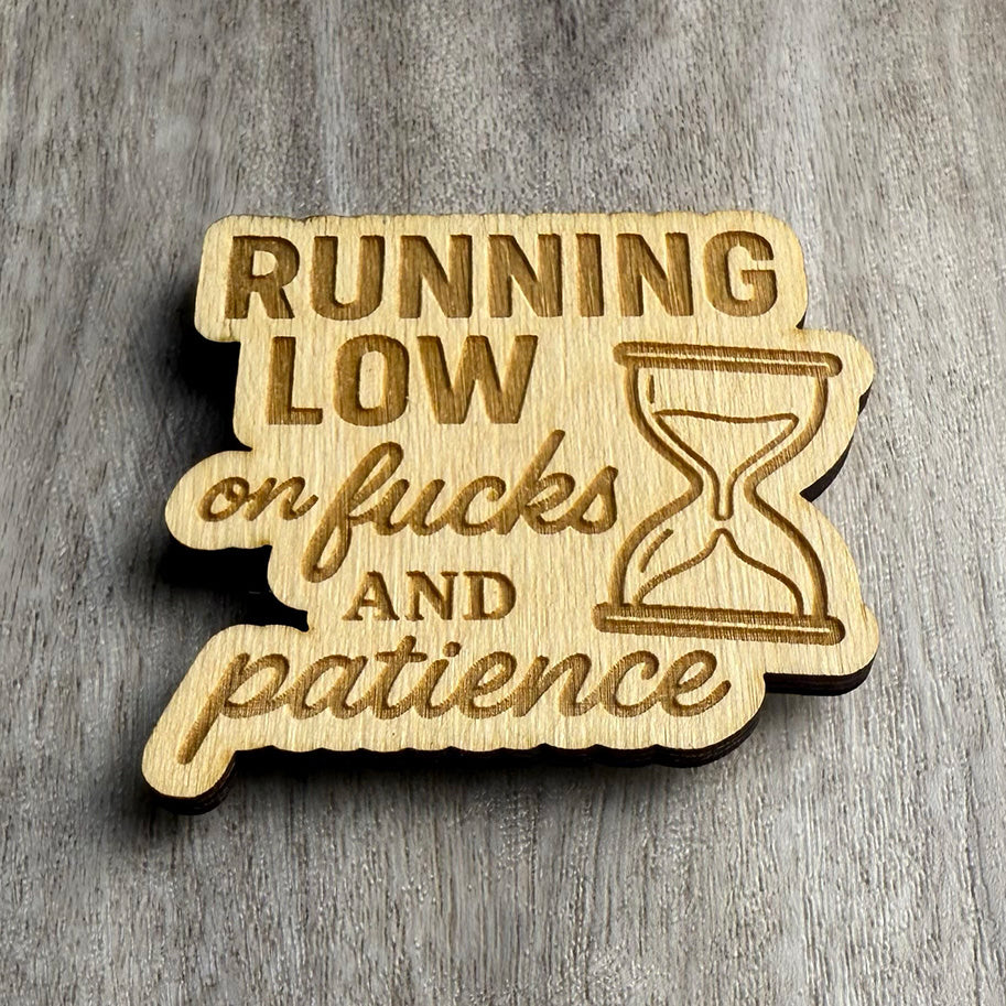 Angled view of a wooden magnet engraved with the text "RUNNING LOW on fucks AND patience" alongside a stylized hourglass illustration. The wood has a scalloped border and sits on a gray-brown wood surface with visible grain texture.