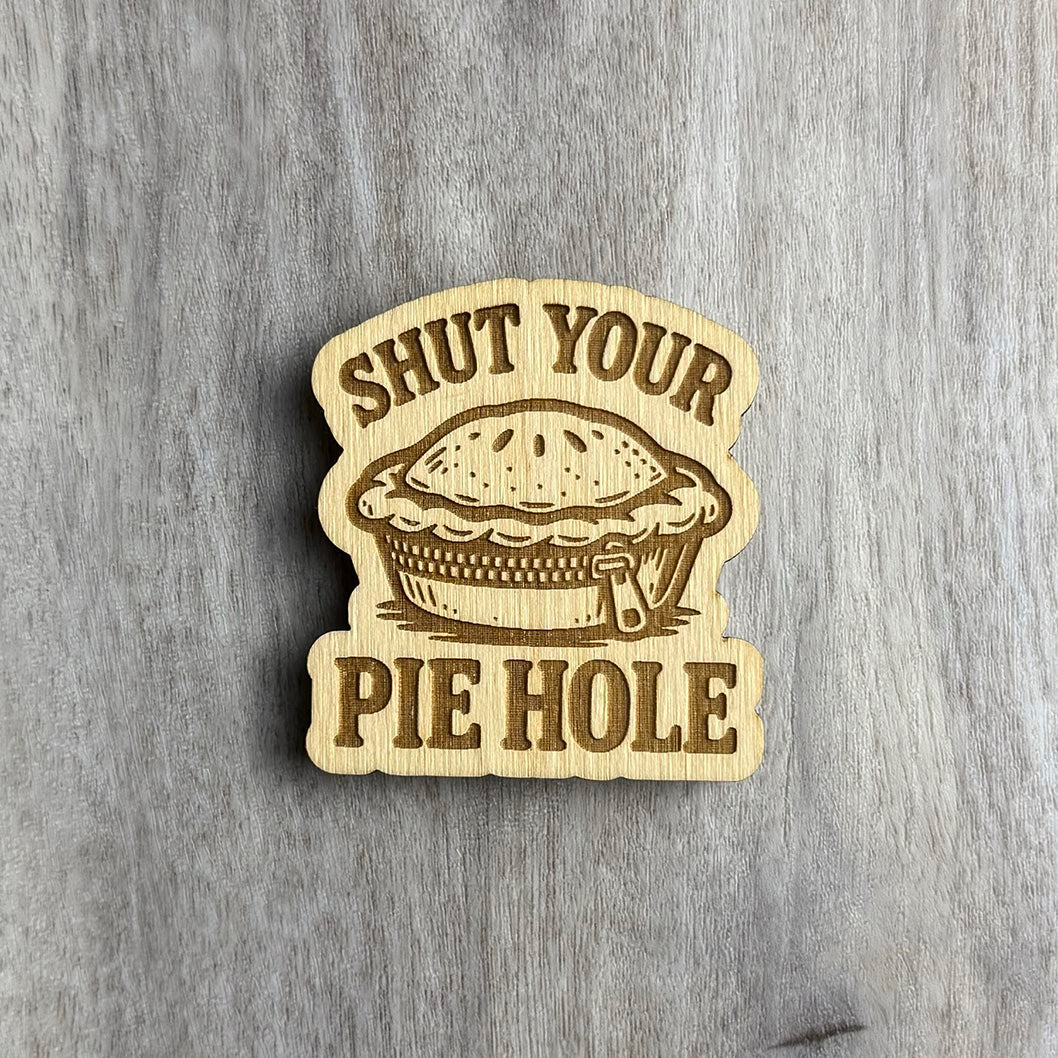Wooden magnet engraved with the phrase "SHUT YOUR PIE HOLE" in bold letters. A detailed illustration of a pie with a zipper across the crust sits in the center, adding humor to the phrase. The magnet has a rounded top and bottom edge with scalloped sides and is photographed on a light wood background.