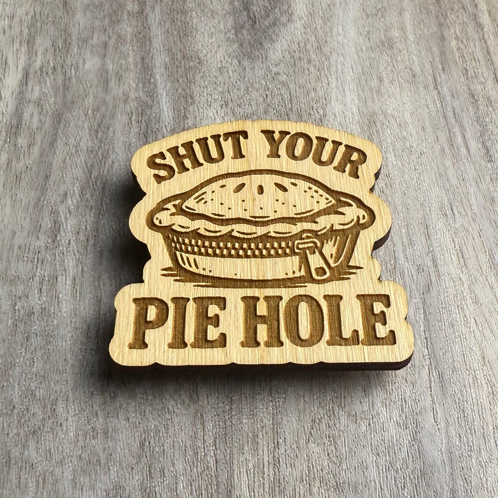 Angled view of a wood magnet engraved with the words "SHUT YOUR PIE HOLE" and a central image of a lidded pie featuring a closed zipper on the crust. The piece has a curved silhouette and is placed on a light gray wood grain surface.