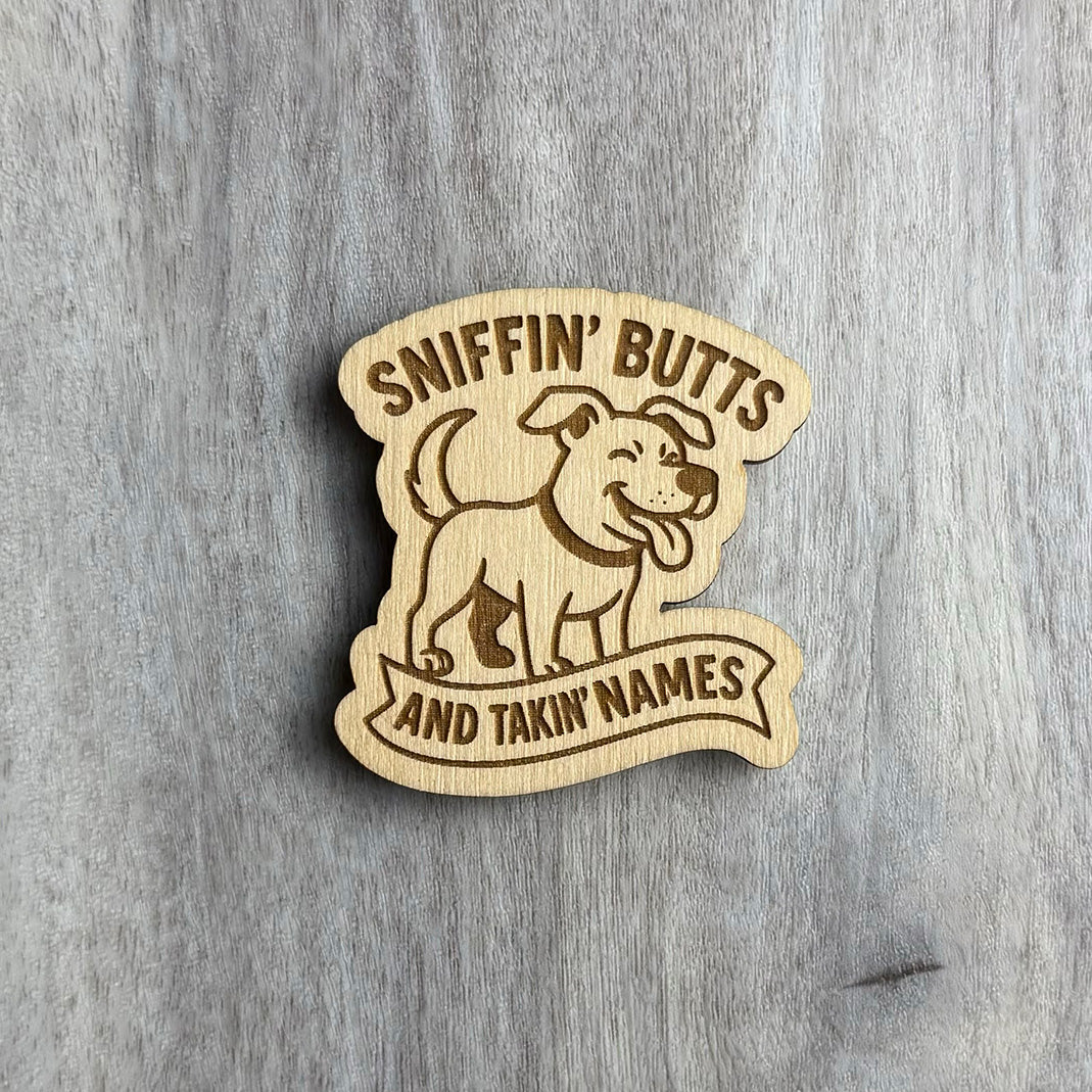 Wooden magnet engraved with the phrase "SNIFFIN' BUTTS AND TAKIN' NAMES" surrounding a playful cartoon dog with a wagging tail and happy expression. The dog stands confidently above the text banner, all carved into a scalloped wood shape resting on a gray wood background.