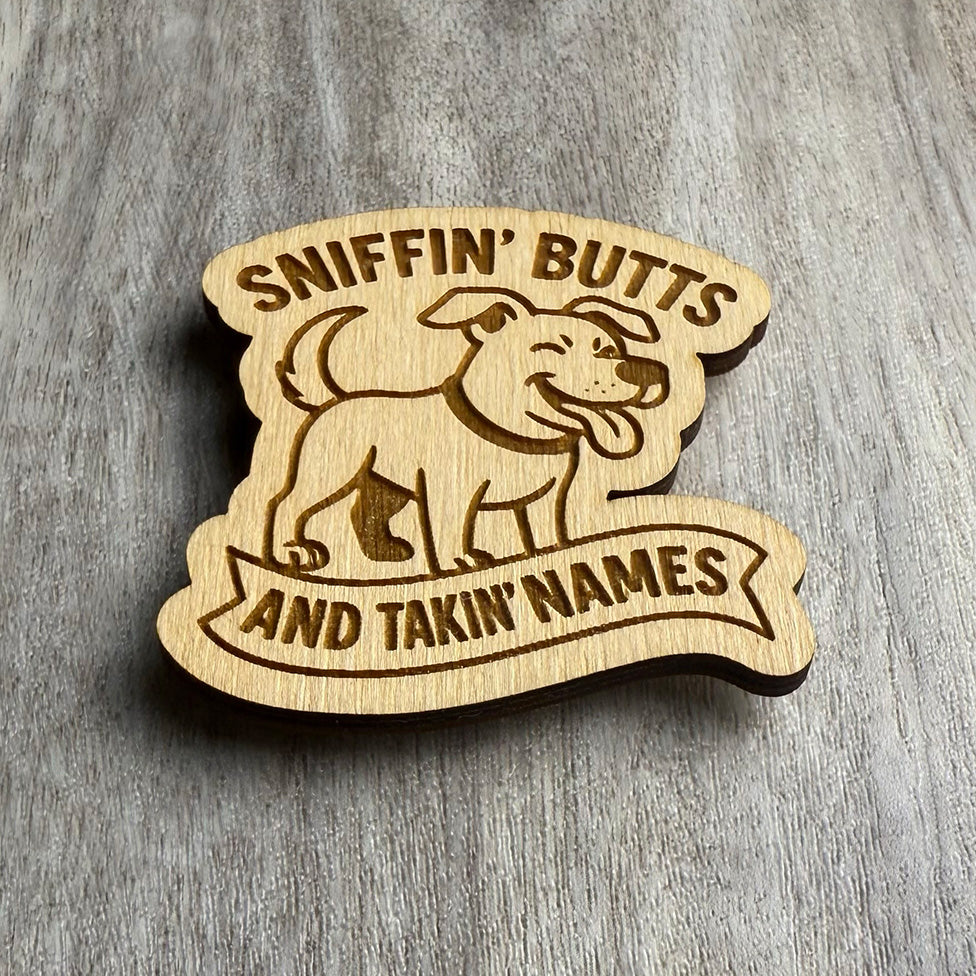 Angled view of a wooden magnet featuring a joyful cartoon dog above the phrase "SNIFFIN' BUTTS AND TAKIN' NAMES." The text is split across the top and bottom, engraved in bold font within a stylized banner. The magnet sits on a textured wood surface with natural grain.
