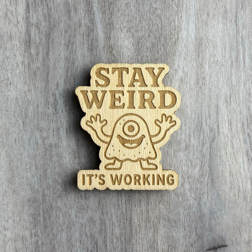 Wooden magnet engraved with the phrase "STAY WEIRD IT'S WORKING" in bold uppercase text. Between the words is a cartoon-style, one-eyed monster with a wide smile and arms raised, carved with playful detail. The background is a lightly textured gray wood surface.