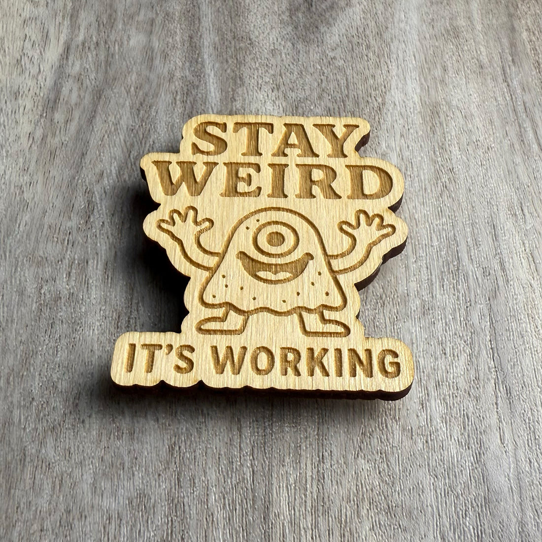 Angled view of a wood magnet featuring the phrase "STAY WEIRD IT'S WORKING" and a central image of a smiling cyclops monster with arms up and happy energy. The engraved design is cut into a wavy-edged shape, resting on a grayish wood background.