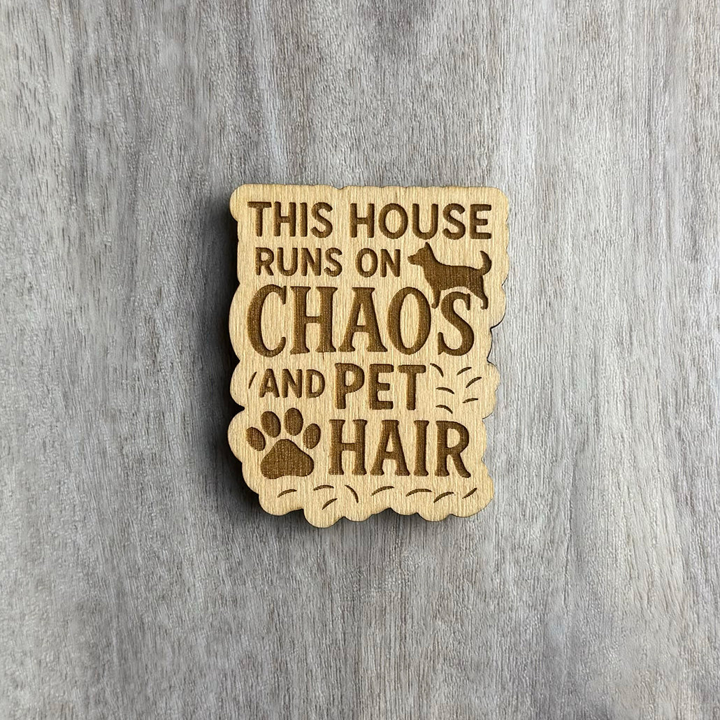 Wooden magnet engraved with the phrase "THIS HOUSE RUNS ON CHAOS AND PET HAIR." The design includes a silhouette of a dog at the top, a large paw print near the bottom, and scattered engraved pet hairs around the text. The border is scalloped and irregular, matching the playful messiness of the message. Background is a light gray wood surface.