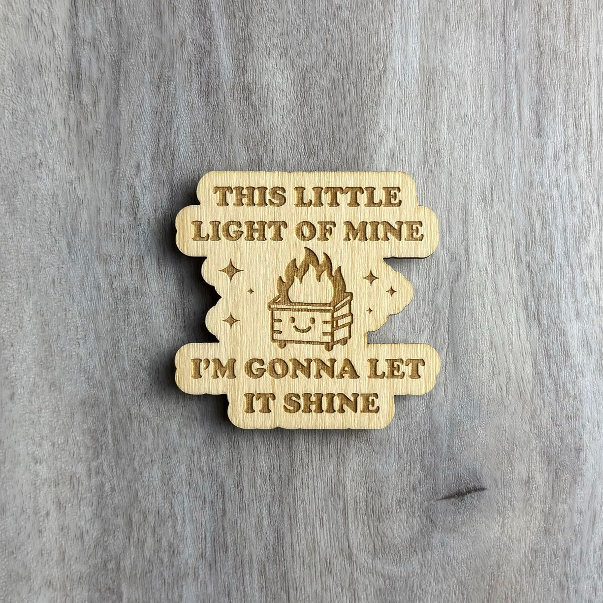 Wooden magnet engraved with the phrase "THIS LITTLE LIGHT OF MINE I'M GONNA LET IT SHINE" in uppercase letters. In the center is a smiling dumpster with flames coming out of it, surrounded by engraved sparkles. The shape has soft, cloud-like curves and is placed on a light gray wood surface.