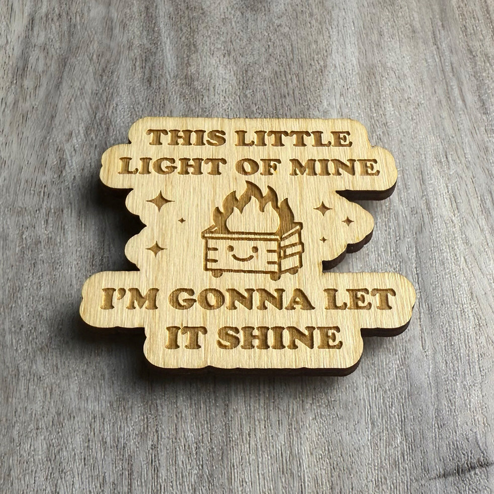 Angled view of a wood magnet featuring the phrase "THIS LITTLE LIGHT OF MINE I'M GONNA LET IT SHINE" and an illustration of a cute, burning dumpster with a cheerful face. Stars and sparkles accent the engraving. The magnet has rounded, blobby edges and sits on a wood-textured background.