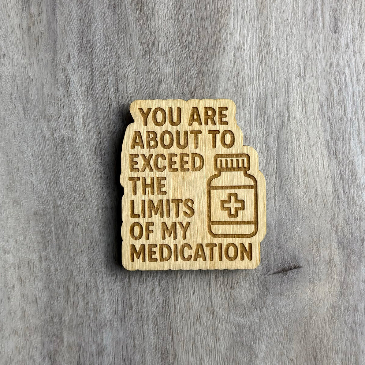 Wooden magnet engraved with the phrase "YOU ARE ABOUT TO EXCEED THE LIMITS OF MY MEDICATION" in bold capital letters. To the right of the text is a simple illustration of a prescription pill bottle with a medical cross symbol. The magnet has a rounded, jagged silhouette and sits on a light wood-grain background.