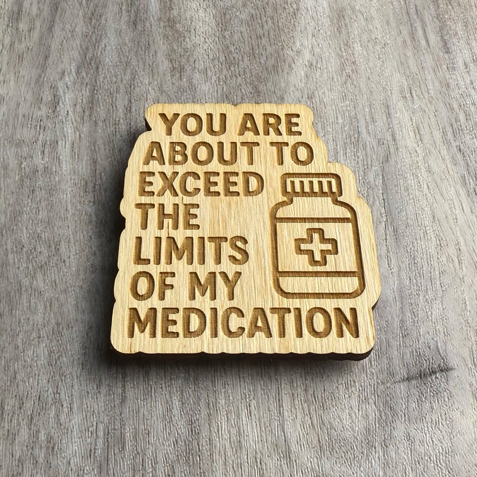 Angled view of a wooden magnet featuring the engraved message "YOU ARE ABOUT TO EXCEED THE LIMITS OF MY MEDICATION" with a pill bottle icon on the right. The engraved design is on a scalloped-edge wood cutout, resting on a textured gray wood surface.